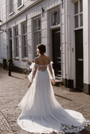 real-bride-review-sophie-rutten-bruidshuis-diana bruid in elegante trouwjurk met off-shoulder mouwen en lange kanten sleep lopend door historische straat.png